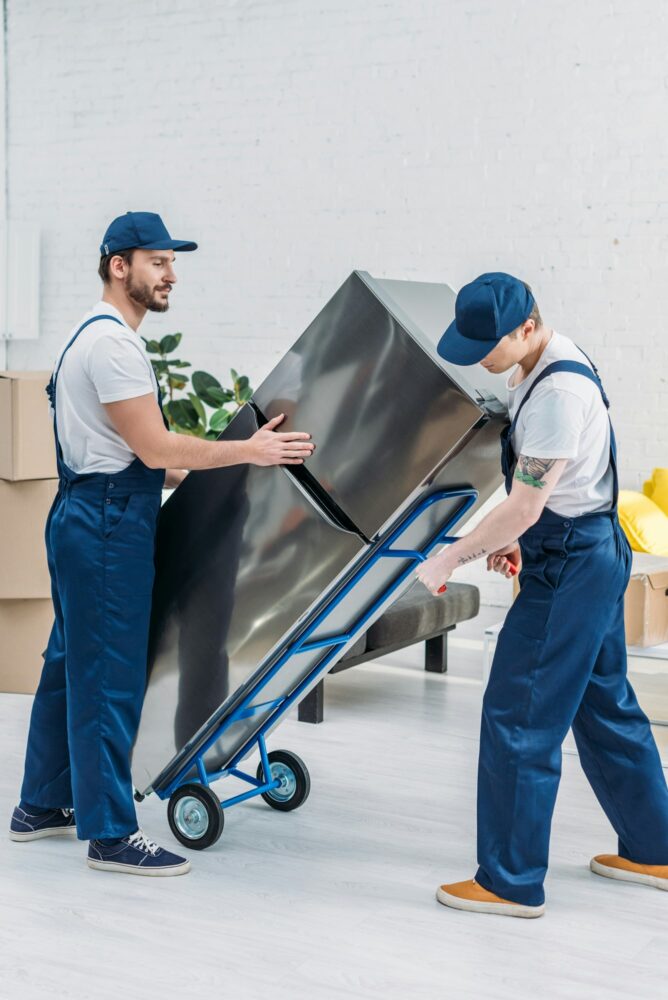 two movers using hand truck while transporting refrigerator in apartment